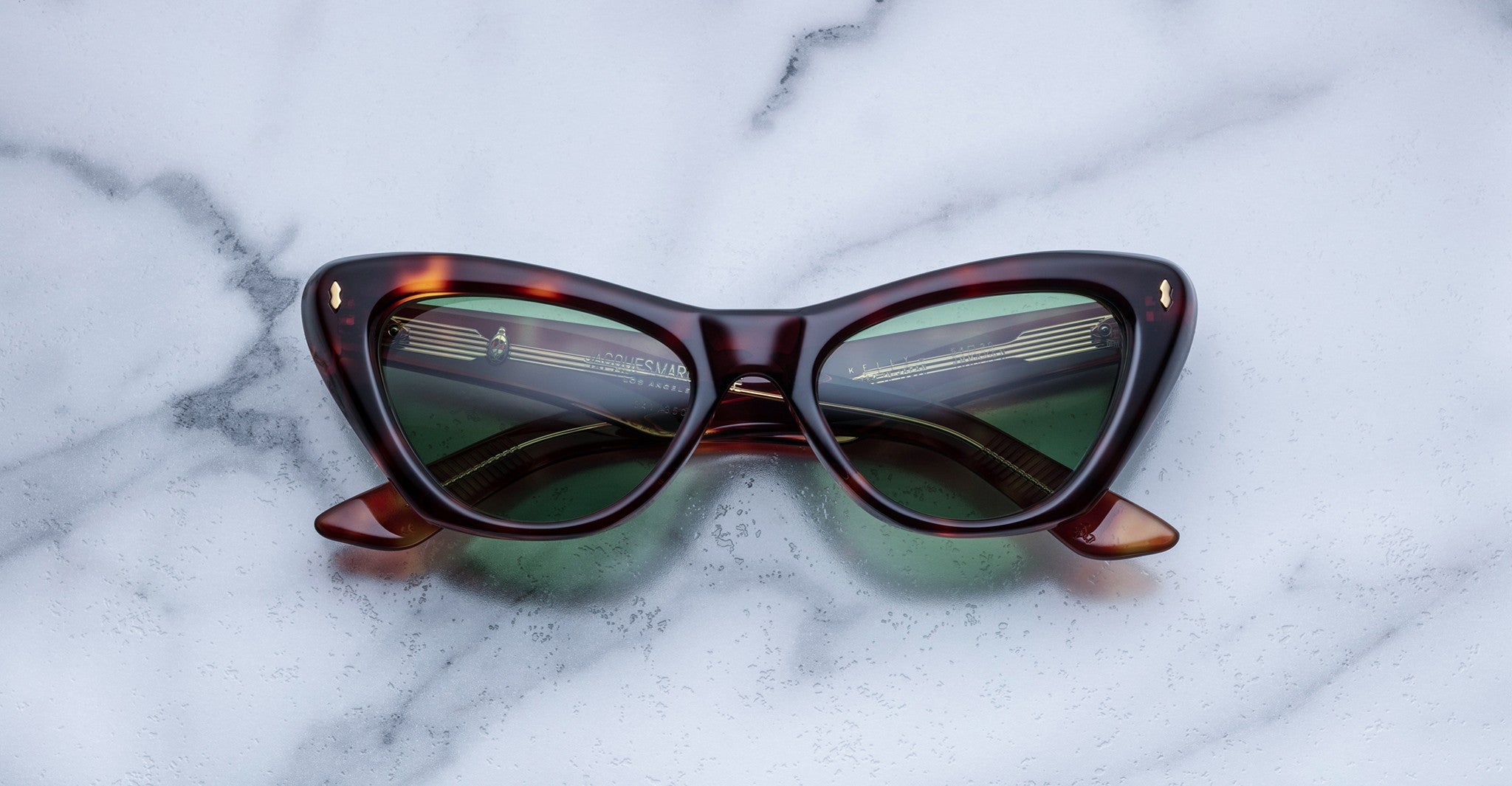 A pair of cat-eye sunglasses with dark green lenses and a brown tortoiseshell frame rests on a light gray marble surface.