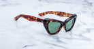 A pair of tortoiseshell cat-eye sunglasses with green lenses is placed on a white marble surface.