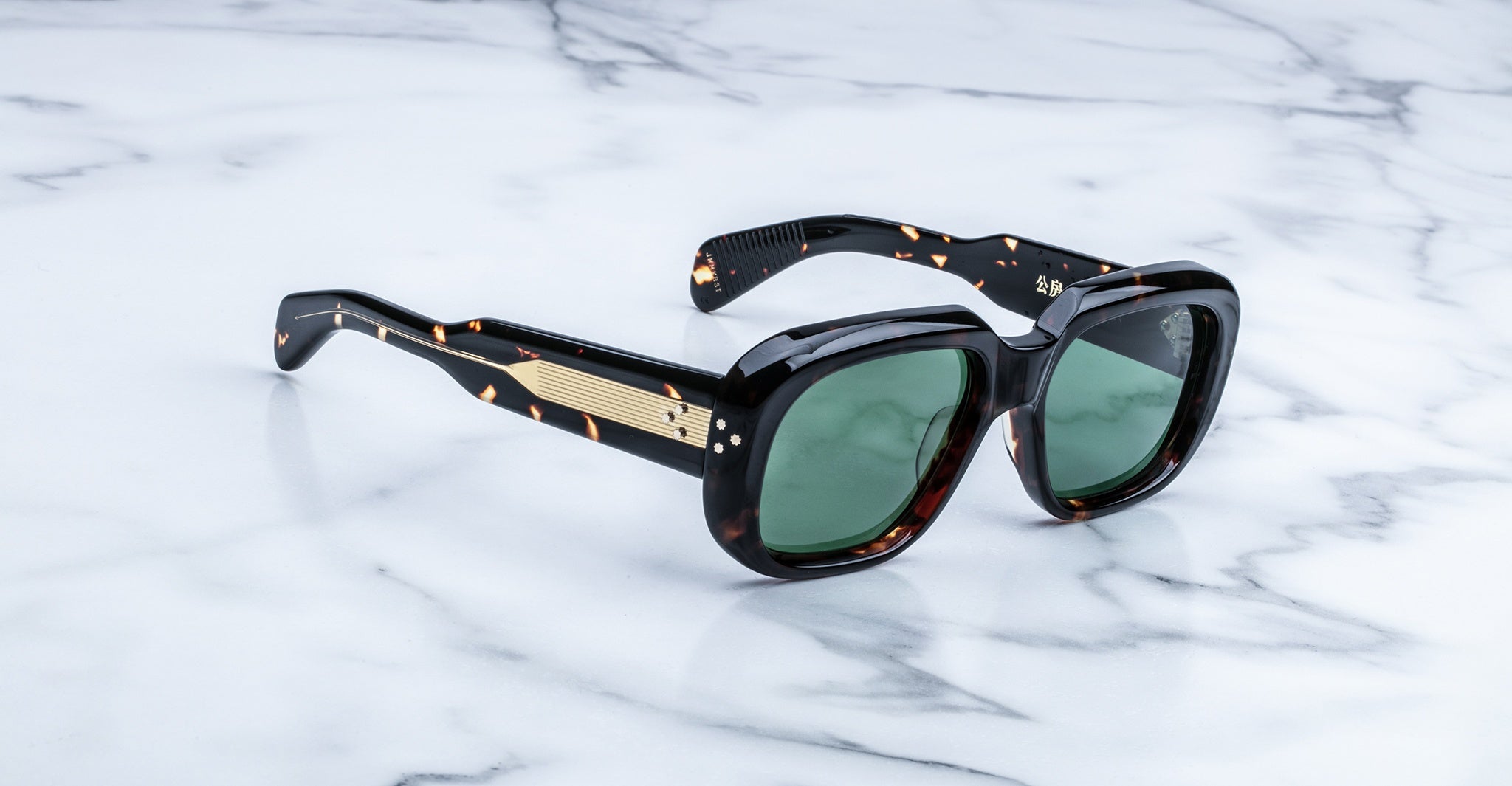 A pair of tortoiseshell sunglasses with thick frames and green lenses, featuring gold accents on the arms, displayed on a white marble surface.
