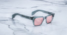 The Jacques Marie Mage Molino sunglasses by COMING SOON feature square, thick, translucent gray frames and pink-tinted lenses, displayed on a white marble surface with gray veining.