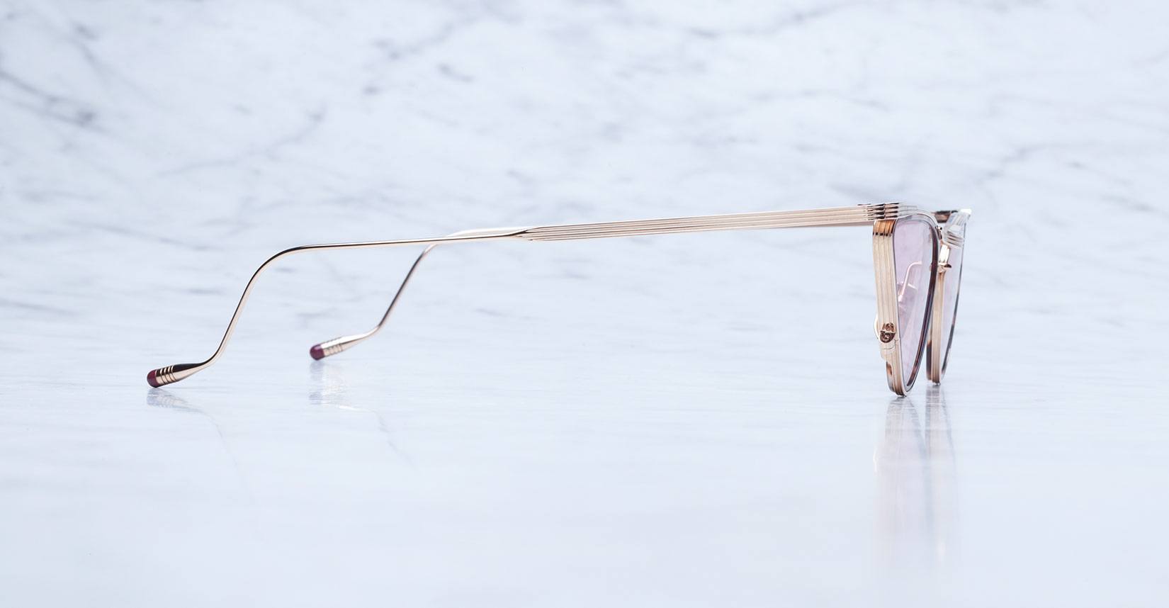 A pair of stylish gold-rimmed eyeglasses shown from the side, resting on a reflective marble surface with a light, cloudy background.