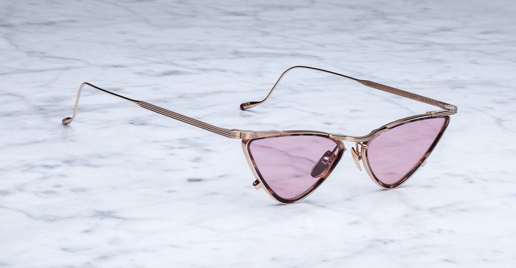 Rose gold cat-eye sunglasses with pink-tinted lenses are placed on a white marble surface. The frames have a modern, angular design and thin metal arms.