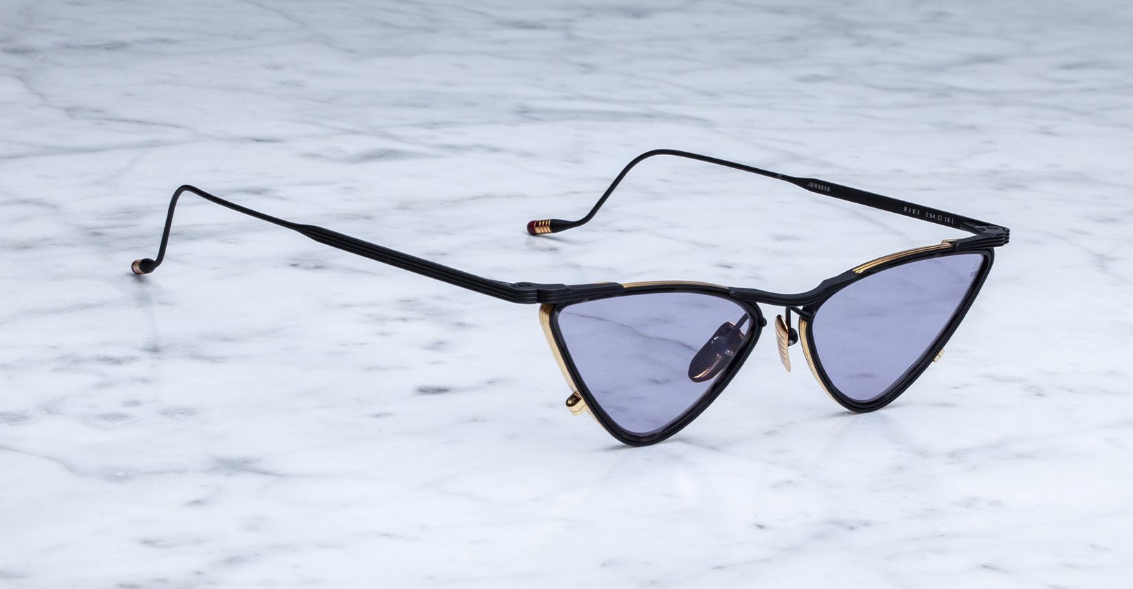 A pair of black and gold cat-eye sunglasses with purple-tinted lenses sits on a light gray marble surface.