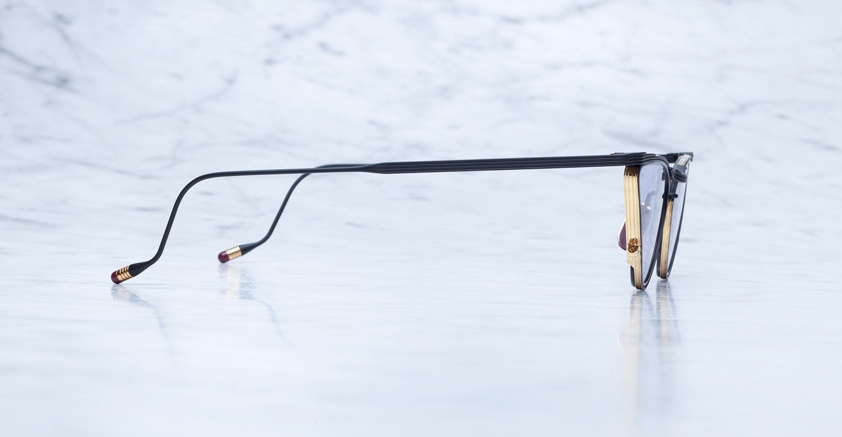 A pair of eyeglasses viewed from the side, showing thin black arms with gold accents on the temple tips, resting on a white marble surface with a light gray background.