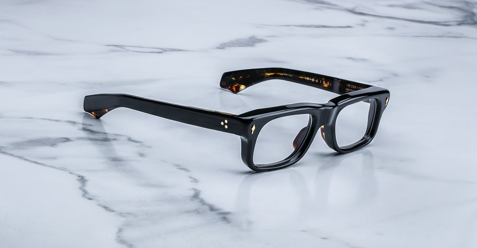 Jacques Marie Mage Saint eyeglasses with thick black rims and gold accents, front view, eyewear placed on a flat surface.