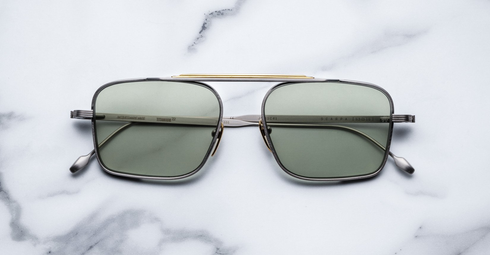 The Jacques Marie Mage Scarpa sunglasses feature green-tinted rectangular lenses, a slim metallic frame, and a gold bar across the top, shown on a white marble surface.