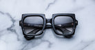 Black square-framed sunglasses with a subtle marbled pattern are placed on a white marble surface, arms crossed, with brand markings visible on the inner temples.