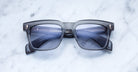 The Jacques Marie Mage Torino sunglasses by Jacques Marie Mage feature bold rectangular black frames with blue tinted lenses, displayed on a white marble surface.