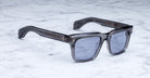 The Jacques Marie Mage Torino, bold rectangular frames in gray translucent acetate with blue-tinted lenses, rest elegantly on a white marble surface.