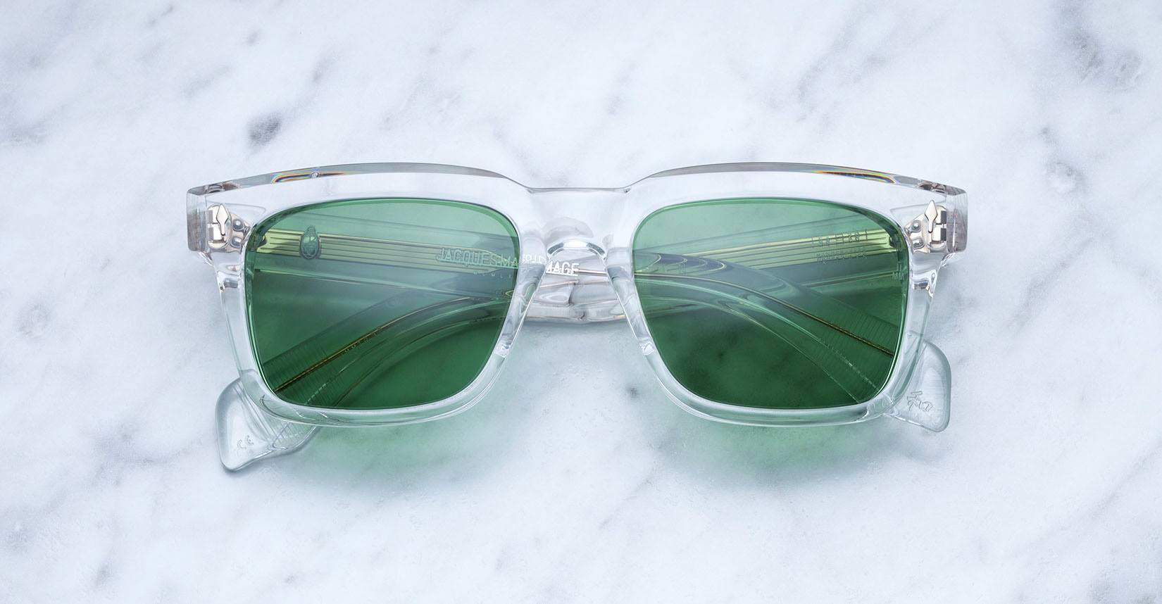 The Jacques Marie Mage Torino sunglasses by Jacques Marie Mage feature a clear frame and green tinted lenses, inspired by bold rectangular eyeglass designs, and rest elegantly on a white marble surface—a tribute to handcrafted acetate eyewear.