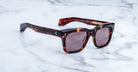 The Jacques Marie Mage Torino sunglasses by Jacques Marie Mage feature tortoiseshell rectangular frames and dark lenses, inspired by bold eyeglass designs, and are displayed elegantly on a white marble surface.