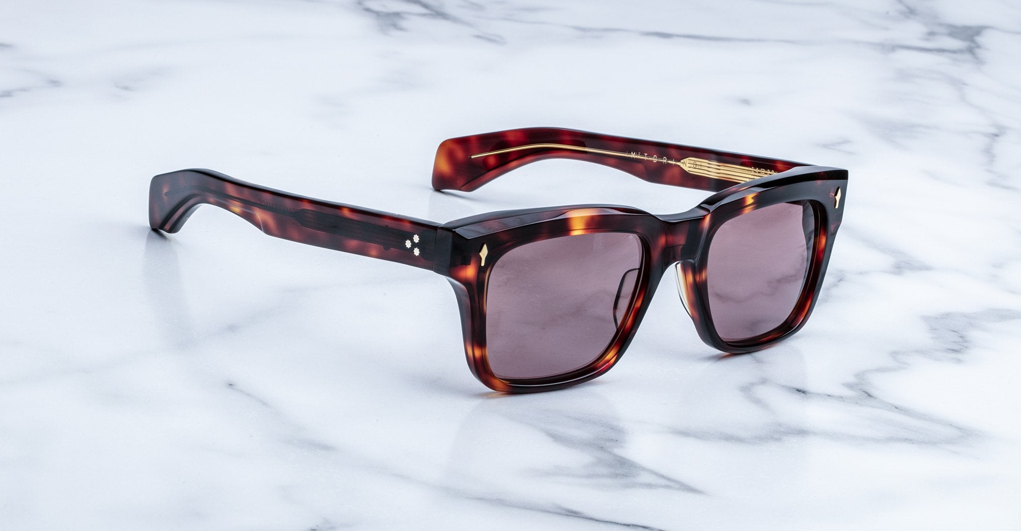 The Jacques Marie Mage Torino sunglasses by Jacques Marie Mage feature tortoiseshell rectangular frames and dark lenses, inspired by bold eyeglass designs, and are displayed elegantly on a white marble surface.