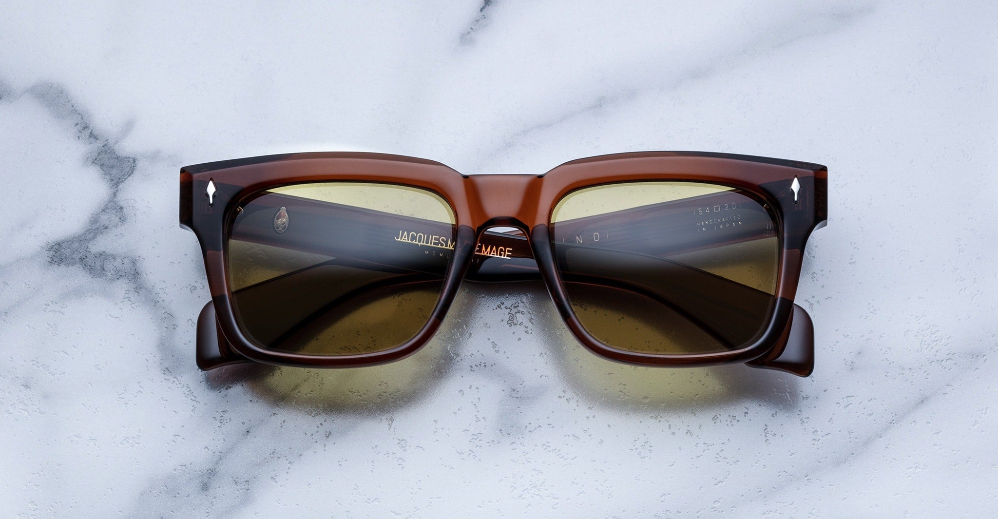 A pair of Jacques Marie Mage Torino rectangular brown sunglasses with dark lenses and bold handcrafted acetate frames rests on a white marble surface.