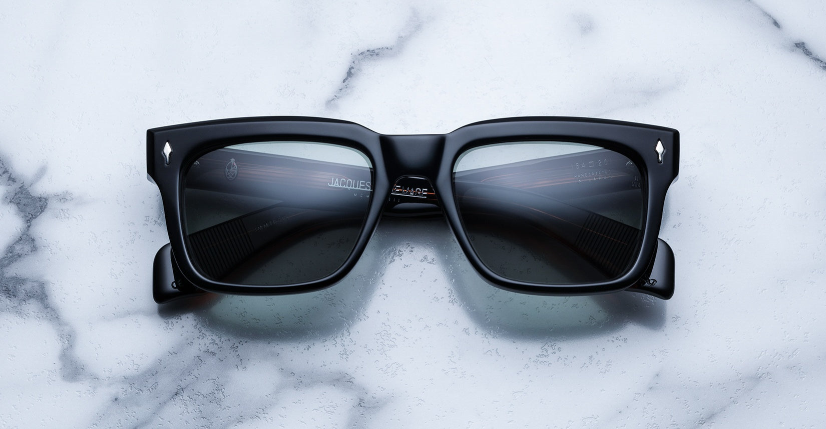 The Jacques Marie Mage Torino, bold black rectangular eyeglass frames with dark lenses, rest on a white marble surface, highlighting the brand's handcrafted acetate artistry.