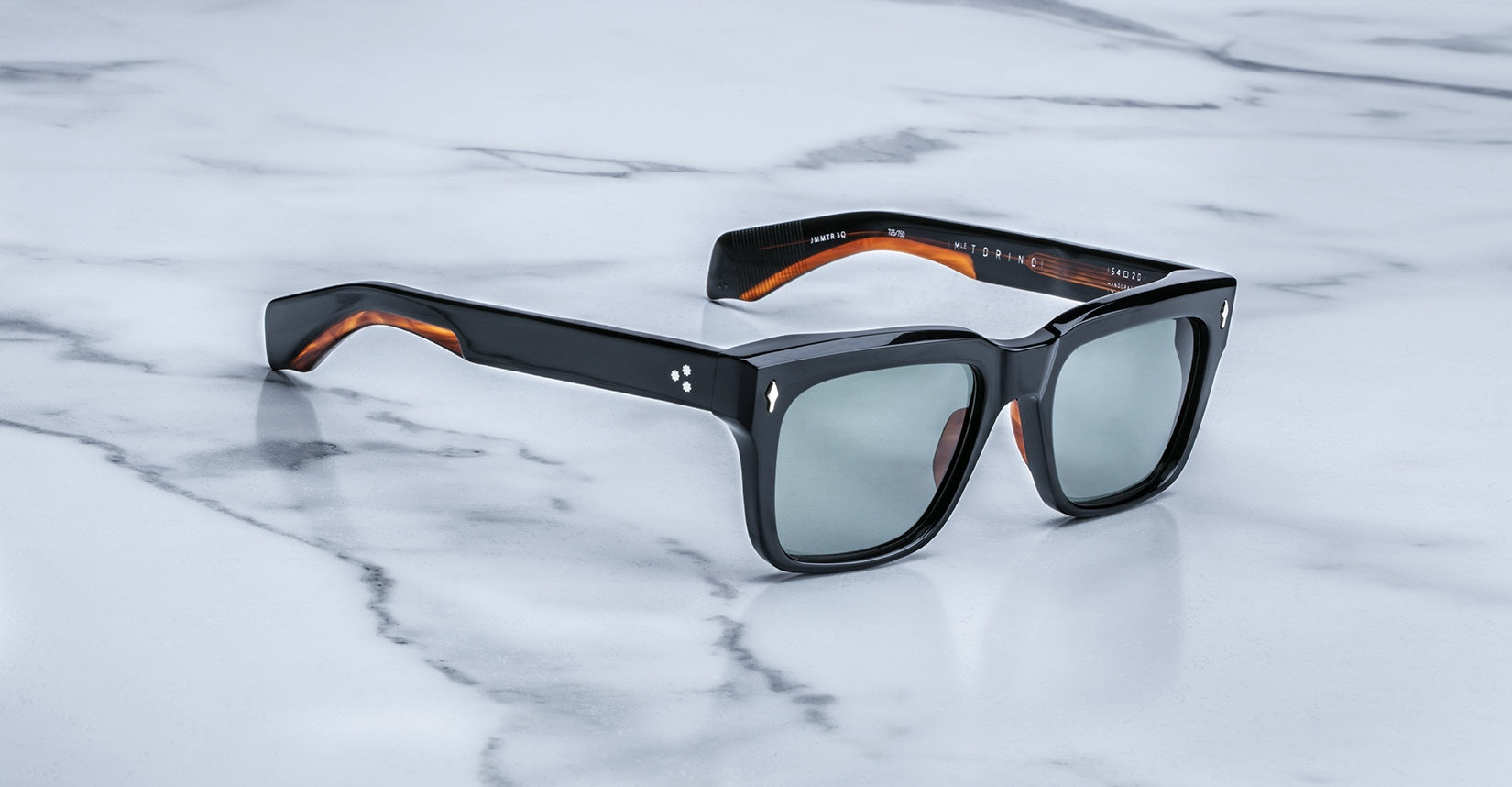 The Jacques Marie Mage Torino sunglasses—black rectangular frames with dark lenses and brown inner arms—crafted from premium acetate, rest stylishly on a white marble surface.