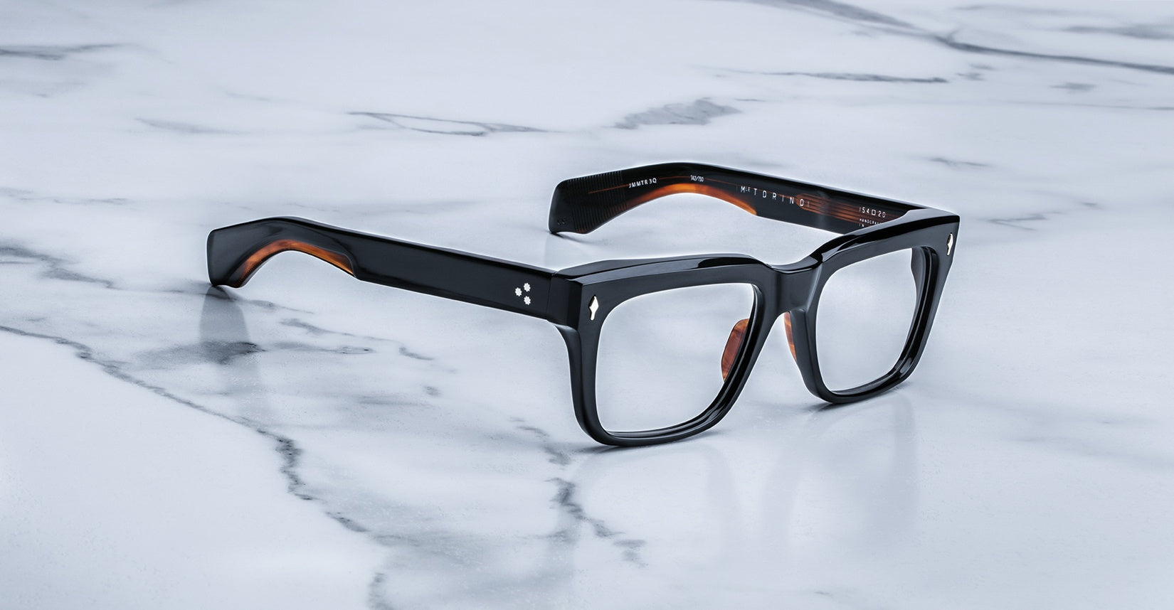 A pair of Jacques Marie Mage Torino rectangular eyeglass frames in black handcrafted acetate by Jacques Marie Mage rests on a white marble surface.