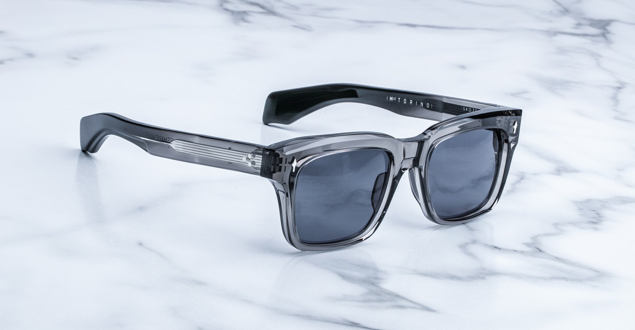 The Jacques Marie Mage Torino sunglasses feature bold rectangular acetate frames and dark square lenses, echoing signature brand style, and are elegantly displayed on a white marble surface.