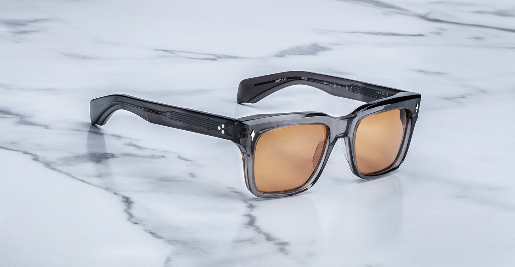 Jacques Marie Mage Torino sunglasses by Jacques Marie Mage feature bold, square dark gray acetate frames and brown-tinted lenses, showcased on a white marble surface—a striking example of handcrafted eyewear.