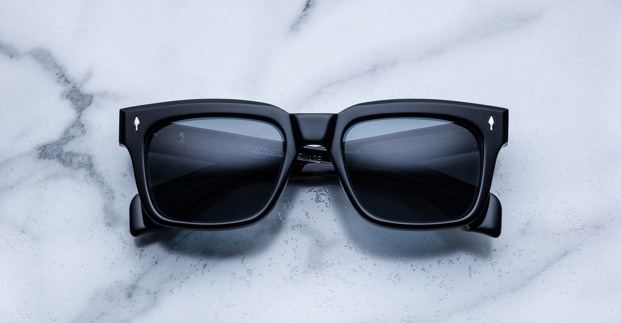 Jacques Marie Mage Torino sunglasses by Jacques Marie Mage feature bold black square frames with dark lenses, inspired by classic rectangular eyeglass styles, showcased on a white marble surface—modern and reminiscent of handcrafted acetate design.