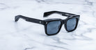 Jacques Marie Mage Torino sunglasses feature bold, black handcrafted acetate frames and dark lenses, resting on a white marble surface. Inspired by iconic style, these Jacques Marie Mage rectangular sunglasses exude timeless elegance.