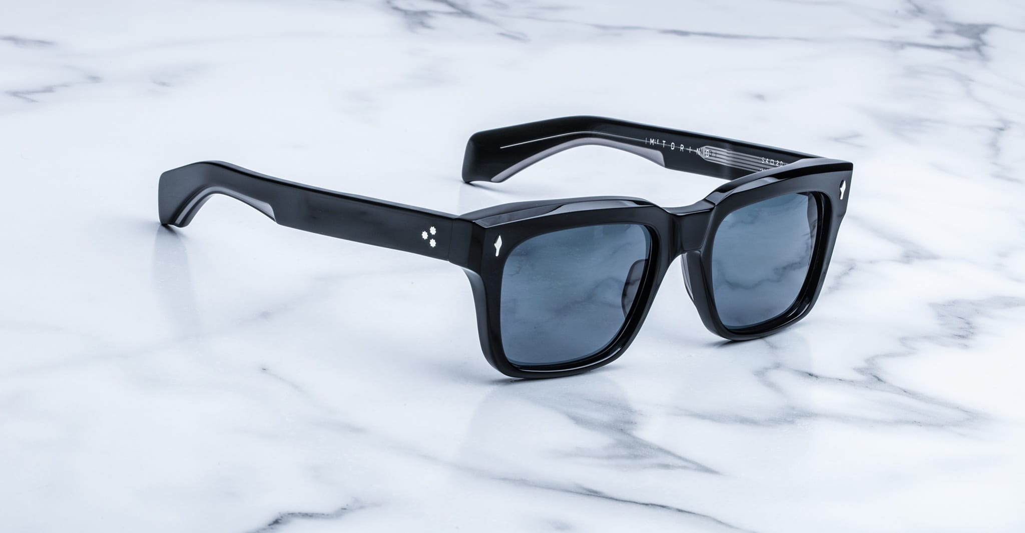 Jacques Marie Mage Torino sunglasses feature bold, black handcrafted acetate frames and dark lenses, resting on a white marble surface. Inspired by iconic style, these Jacques Marie Mage rectangular sunglasses exude timeless elegance.