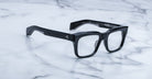 A pair of Jacques Marie Mage Torino eyeglass frames by Jacques Marie Mage sit on a white marble surface, highlighting the brand's handcrafted acetate and bold rectangular design.