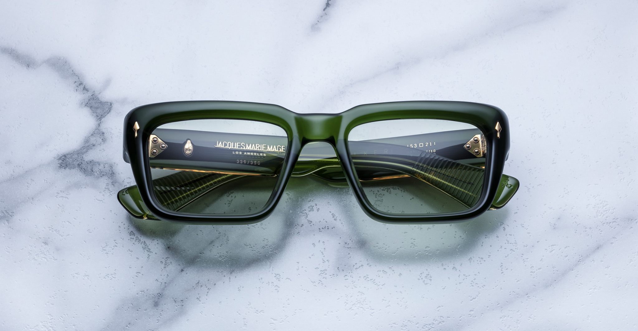 Green rectangular sunglasses with thick frames are placed on a white marble surface. The lenses reflect the image of a stairway with golden railings, adding an artistic effect.