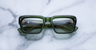 Green rectangular sunglasses with thick frames are placed on a white marble surface. The lenses reflect the image of a stairway with golden railings, adding an artistic effect.