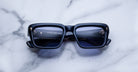 Black rectangular sunglasses with blue-tinted lenses rest on a white marble surface. The lenses reflect a modern interior scene with text and geometric shapes visible in the reflection.