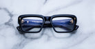 Thick black rectangular eyeglasses are placed face up on a white marble surface, with blue details and reflections visible on the lenses and inside the frame arms.