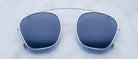 The Jacques Marie Mage Zephirin 47 Clip On features blue-tinted clip-on lenses with a silver metal frame and bridge, displayed on a light marble-textured background.