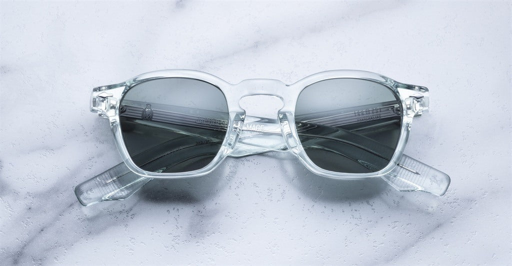 Jacques Marie Mage Zephirin sunglasses with clear frames and dark lenses rest on white marble—a sophisticated tribute to limited edition eyewear by Jacques Marie Mage, beautifully crafted from premium acetate.