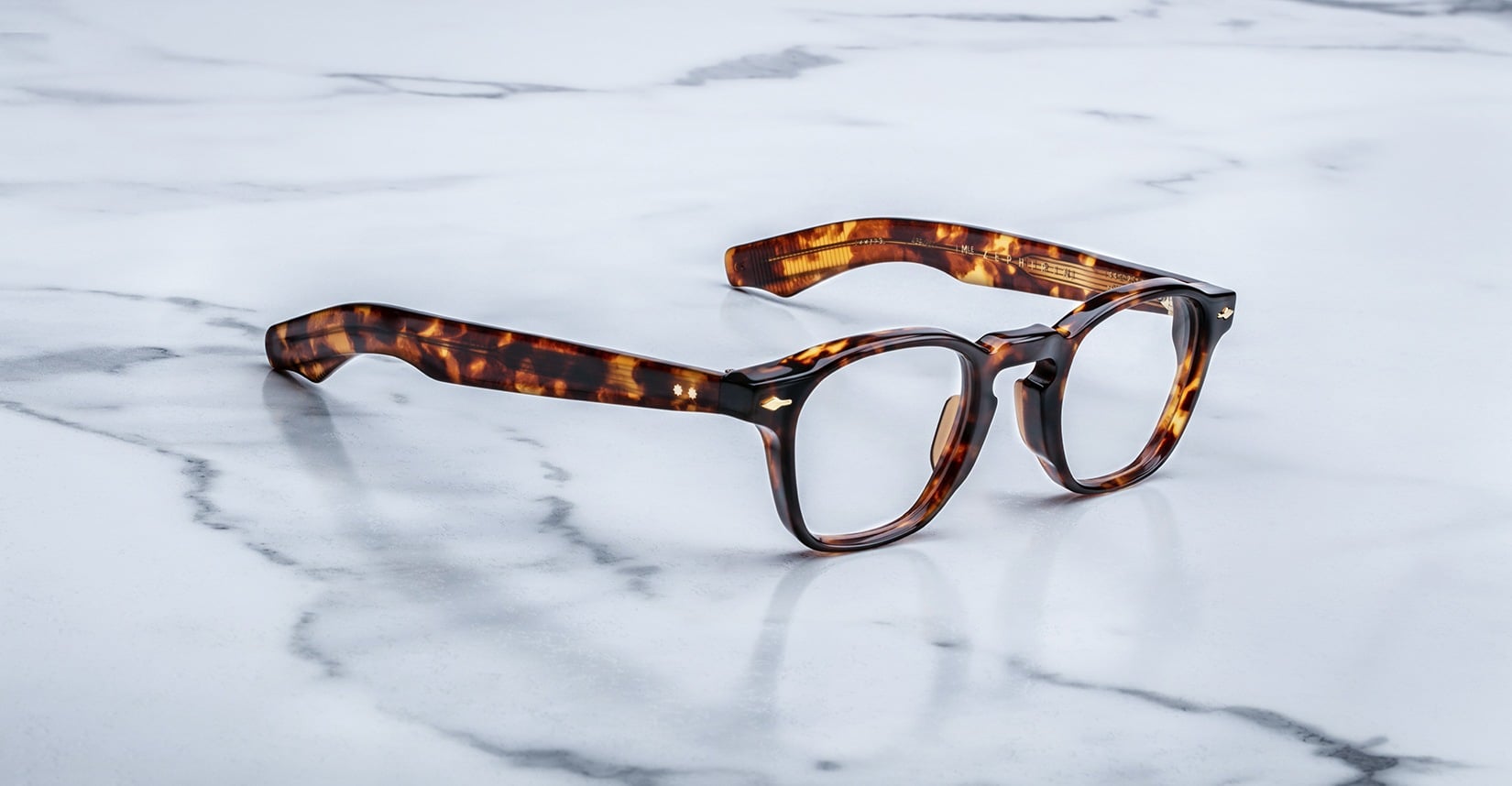 A pair of Jacques Marie Mage Zephirin handcrafted acetate spectacles with round lenses rests on a white marble surface.