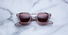 Handcrafted Jacques Marie Mage Zephirin sunglasses, featuring clear acetate frames and pink tinted lenses, rest on white marble—a tribute to the brand’s iconic limited edition eyewear.