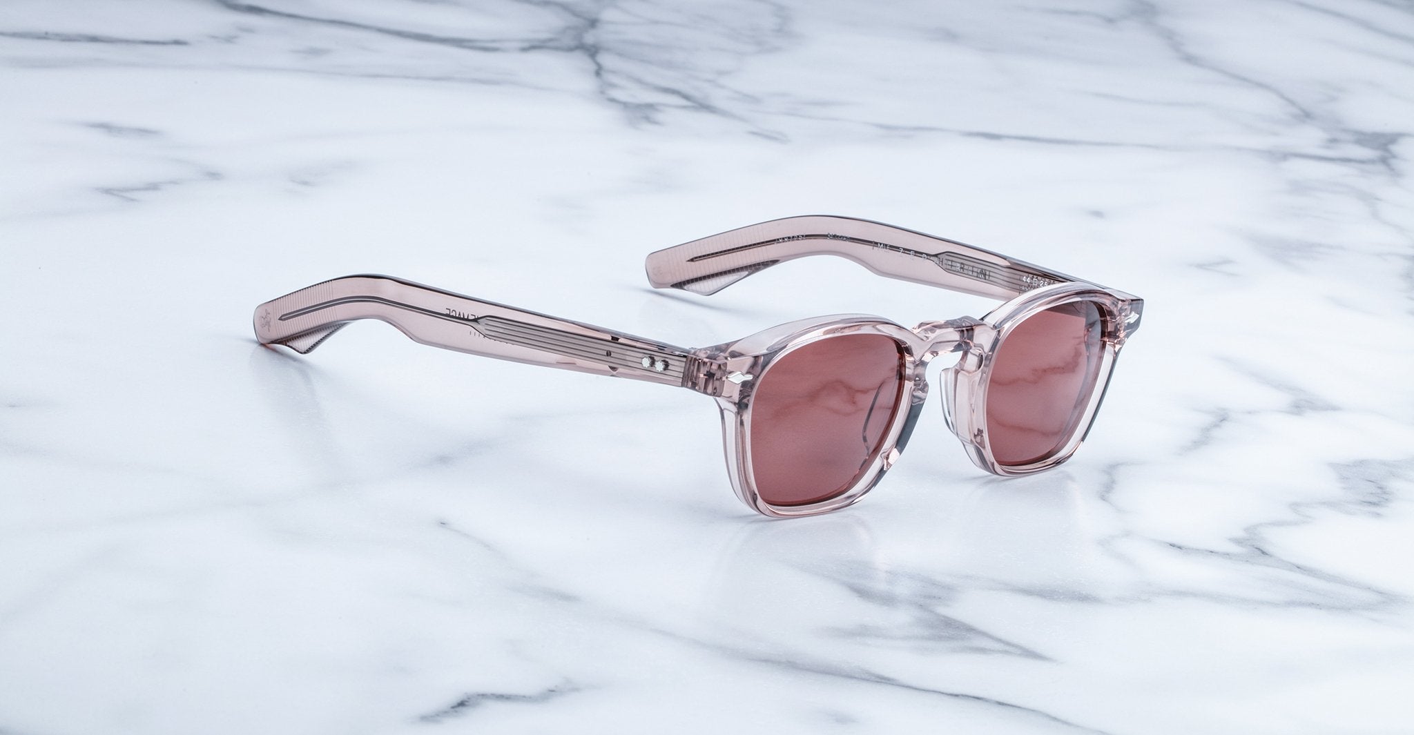 The Jacques Marie Mage Zephirin, limited edition sunglasses with transparent pink acetate frames, rest on a white marble surface, highlighting the brand’s handcrafted elegance.