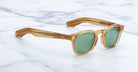 A pair of Jacques Marie Mage Zephirin sunglasses featuring amber square acetate frames and green lenses, displayed on a white marble surface.
