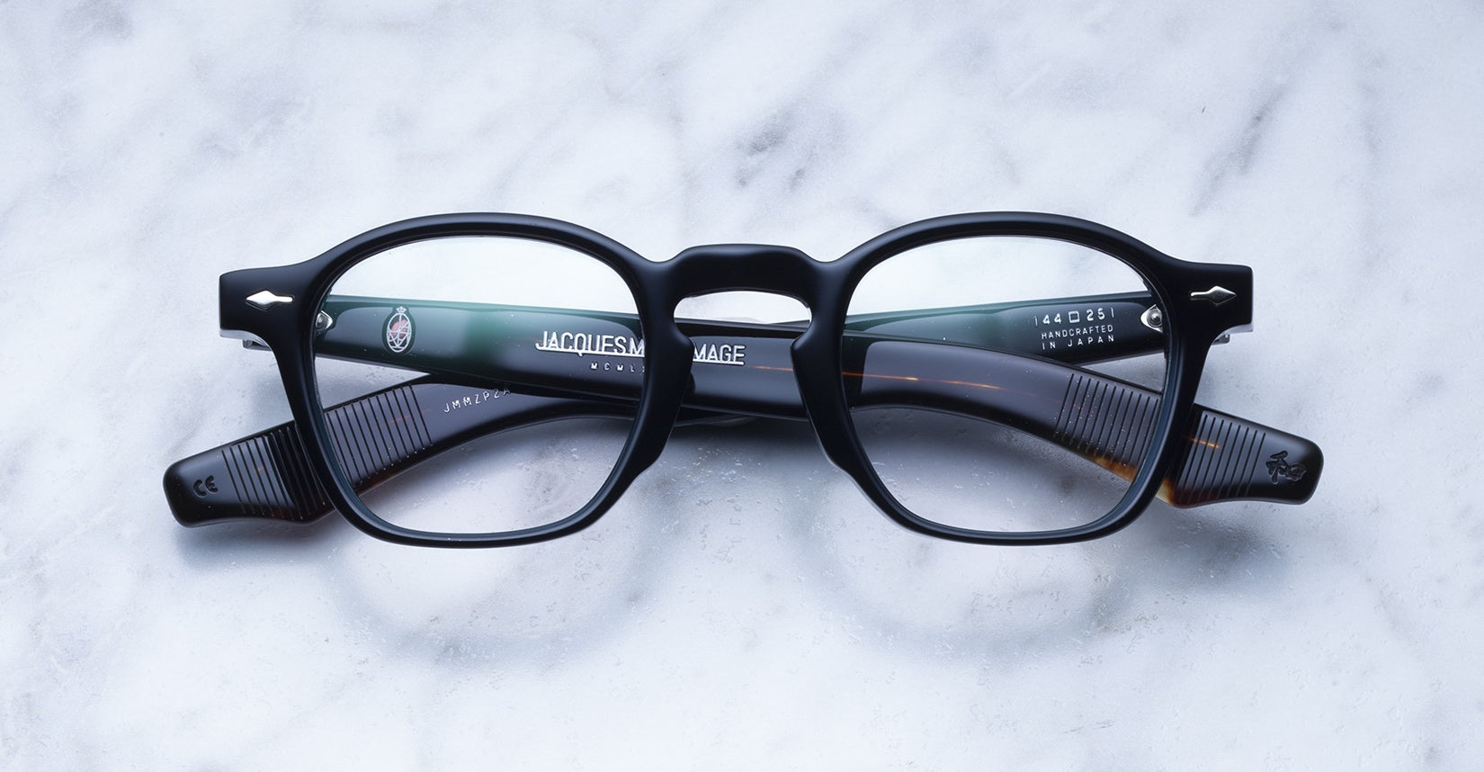 A pair of Jacques Marie Mage Zephirin black round eyeglasses with thick handcrafted acetate frames and ribbed temple arms rests on a white marble surface.