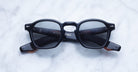 A pair of Jacques Marie Mage Zephirin black acetate sunglasses with dark lenses from Jacques Marie Mage is displayed on a white marble surface.
