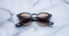 A pair of Jacques Marie Mage Zephirin sunglasses with brown lenses and a dark frame sits on white marble, highlighting the exclusive appeal of this limited edition by Jacques Marie Mage.