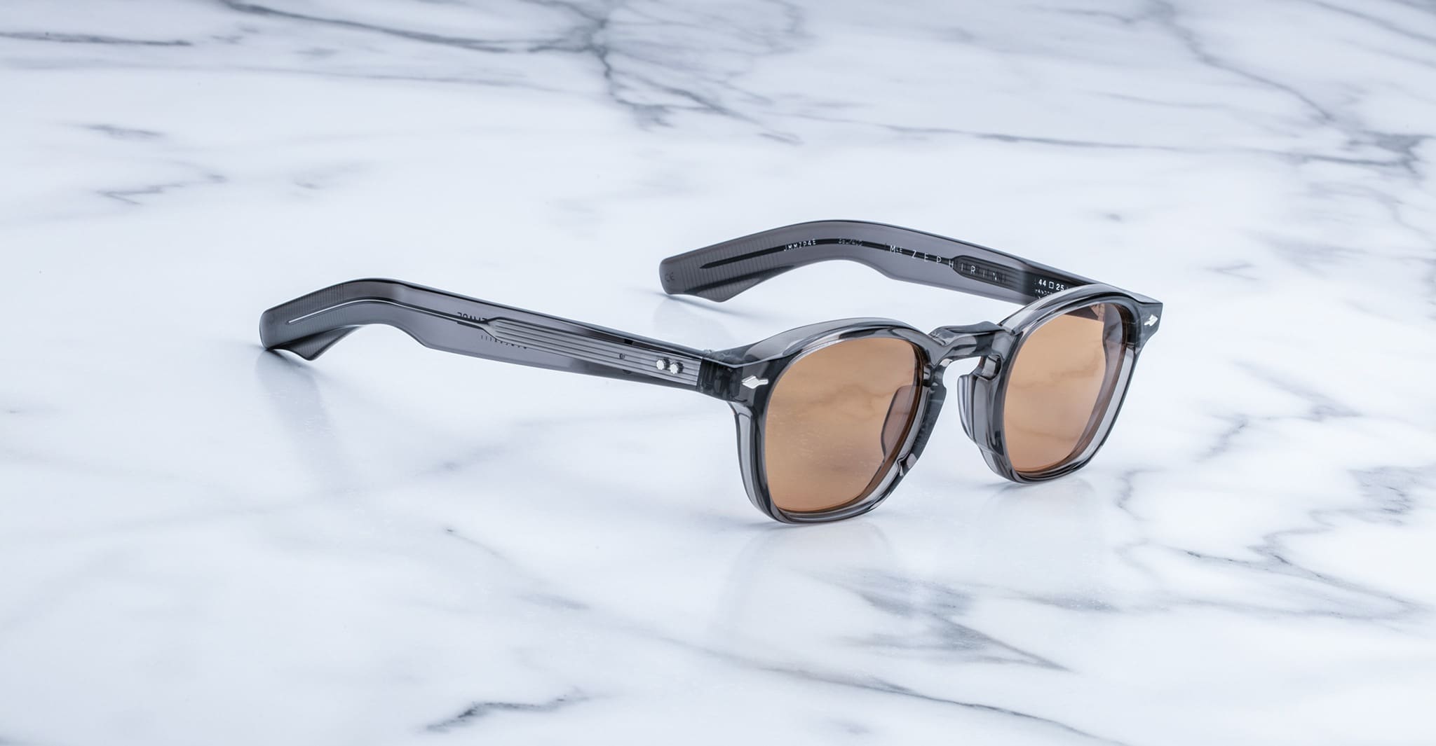The Jacques Marie Mage Zephirin sunglasses, featuring brown lenses and thick translucent gray frames, are displayed elegantly on a white marble surface.