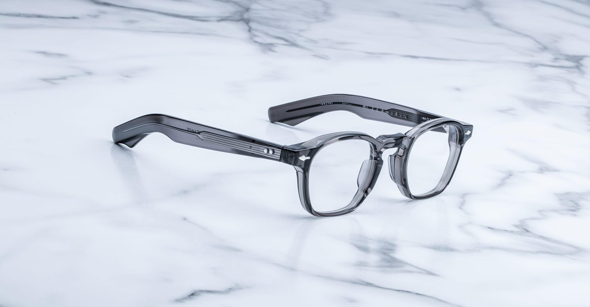 The Jacques Marie Mage Zephirin, transparent gray handcrafted acetate spectacles with thick frames, rest on a white marble surface.