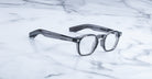 The Jacques Marie Mage Zephirin, transparent gray handcrafted acetate spectacles with thick frames, rest on a white marble surface.