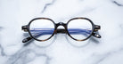 The Jacques Marie Mage Clark are round tortoiseshell eyeglasses with blue-tinted lenses, inspired by midcentury design, shown on a white marble surface.