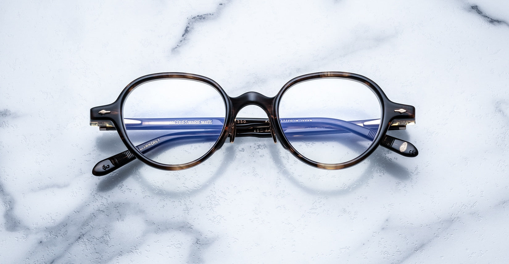 The Jacques Marie Mage Clark are round tortoiseshell eyeglasses with blue-tinted lenses, inspired by midcentury design, shown on a white marble surface.