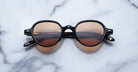 The Jacques Marie Mage Clark sunglasses feature round, black frames and brown-tinted lenses, showcasing midcentury-inspired style as they rest elegantly on a white marble surface.
