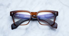 The Jacques Marie Mage Molino eyeglasses by COMING SOON feature thick, rectangular brown and translucent purple frames. Resting on a white marble surface, the temples are neatly folded beneath the frames.