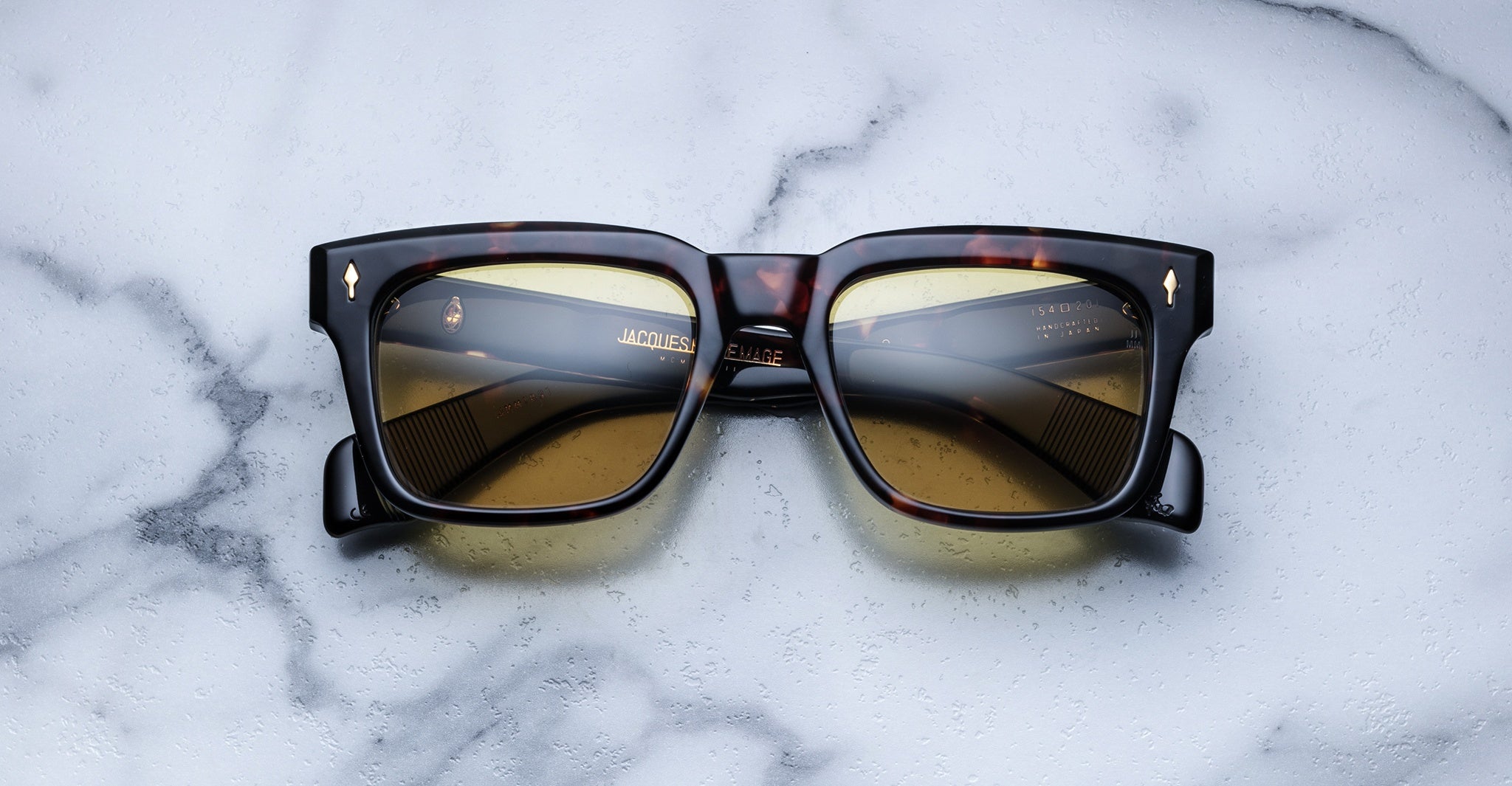 The Jacques Marie Mage Torino, bold rectangular eyeglass frames in tortoiseshell with brown lenses by Jacques Marie Mage, rest on a white marble surface, evoking the essence of handcrafted acetate eyewear.