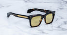 A pair of Jacques Marie Mage Torino rectangular sunglasses with yellow lenses and gold arm detailing, handcrafted from acetate, rest on a white marble surface.