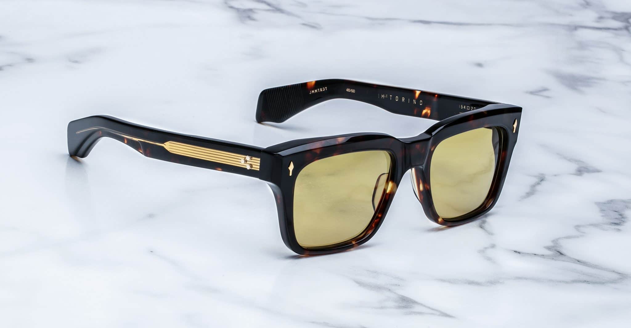 A pair of Jacques Marie Mage Torino rectangular sunglasses with yellow lenses and gold arm detailing, handcrafted from acetate, rest on a white marble surface.