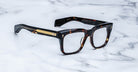 Jacques Marie Mage Torino eyeglasses: bold, handcrafted acetate frames in tortoiseshell with gold temple detailing, inspired by the iconic Jacques Marie Mage brand, elegantly displayed on a white marble surface.